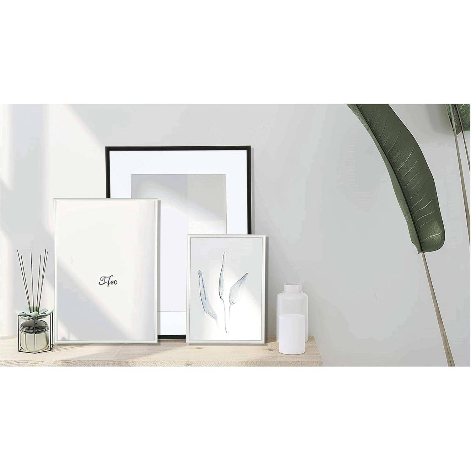 Drei minimalistische Rahmen auf einem Holzregal: ein großer Rahmen mit 'Baum', ein mittlerer Rahmen mit geometrischen Formen und ein kleiner pflanzenmotivierter Rahmen, neben einer Pflanze und einer weißen Flasche.