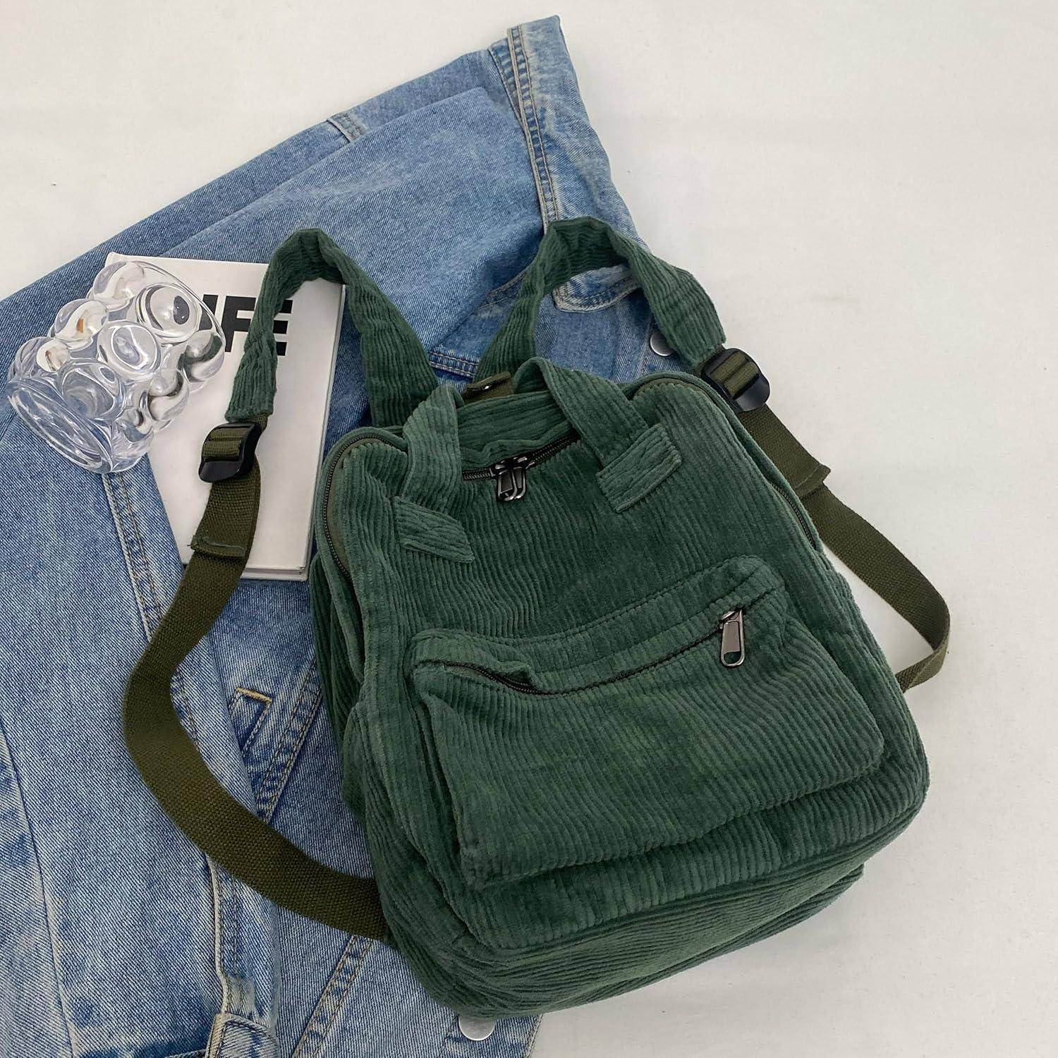 Ein grüner Cordruck-Rucksack mit Vordertasche und verstellbaren Trägern liegt auf gefalteten Bluejeans neben einem durchsichtigen Glas, das kleine Ringe enthält.
