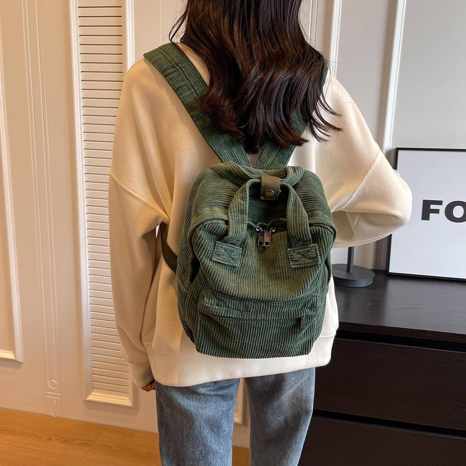 Person mit langen Haaren in einem cremefarbenen Pullover und Jeans steht drinnen, dem Wandzugewandt, und trägt einen grünen Cordage-Rucksack.