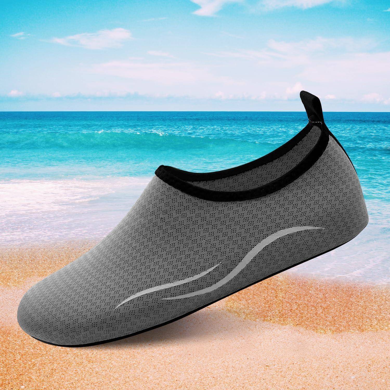 Ein einzelner Wasserschuh in Grau mit schwarzer Einfassung, der auf einem sandigen Strand mit dem Ozean und blauem Himmel im Hintergrund positioniert ist.