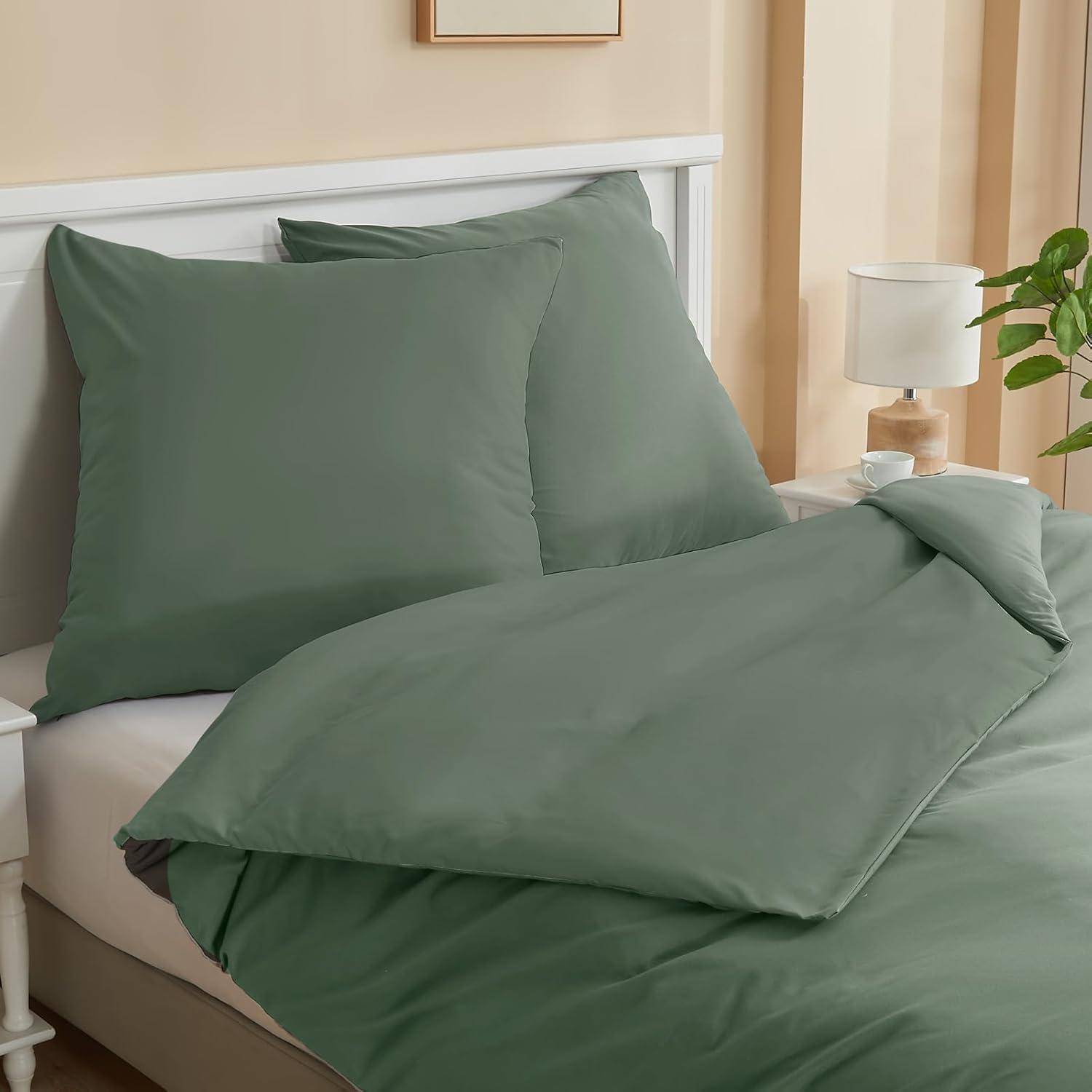 Ein sorgfältig gemachtes Bett mit salbeigrüner Bettwäsche, zwei Kissen, ein Nachttisch mit einer Lampe und eine kleine Pflanze daneben in einer gemütlichen Schlafzimmeratmosphäre.