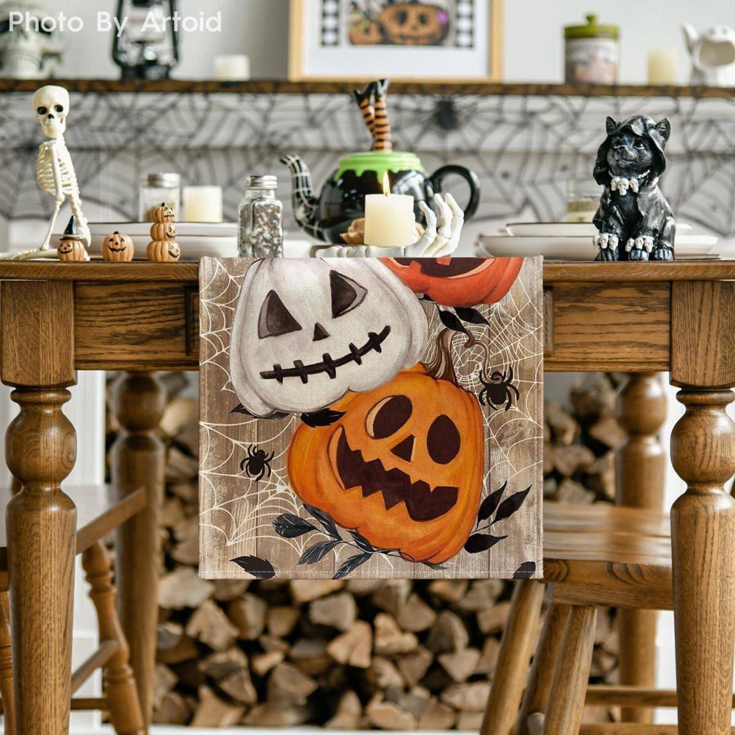 Ein rustikaler Tisch, geschmückt mit Halloween-Motiven wie Kürbislampen, einer Schwarze-Katze-Figur und einem Skelett. Ein Handtuch mit Kürbismotiv hängt vom Tisch.