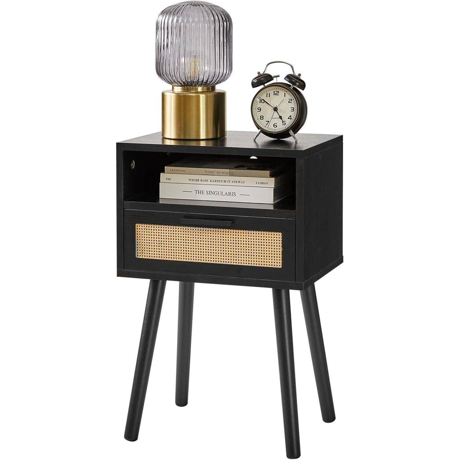 Ein schwarzer Nachttisch mit einer geflochtenen Schublade, in der Bücher und eine Uhr liegen; eine Lampe und ein kleiner Wecker stehen auf der Oberfläche.
