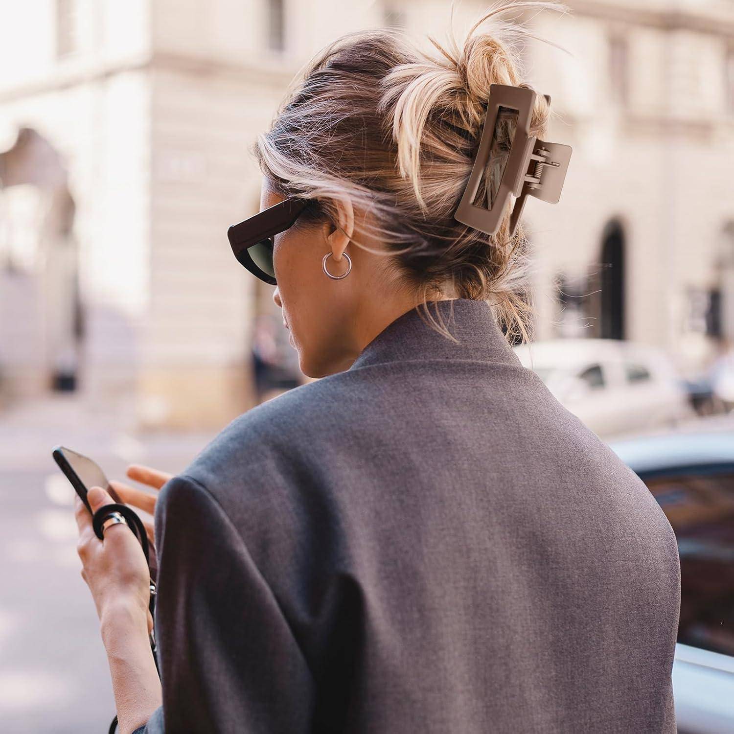 Eine Person mit Sonnenbrille und Haarspange geht in einer Stadtumgebung und schaut auf ein Smartphone. Im Hintergrund sind verschwommene Gebäude zu sehen.