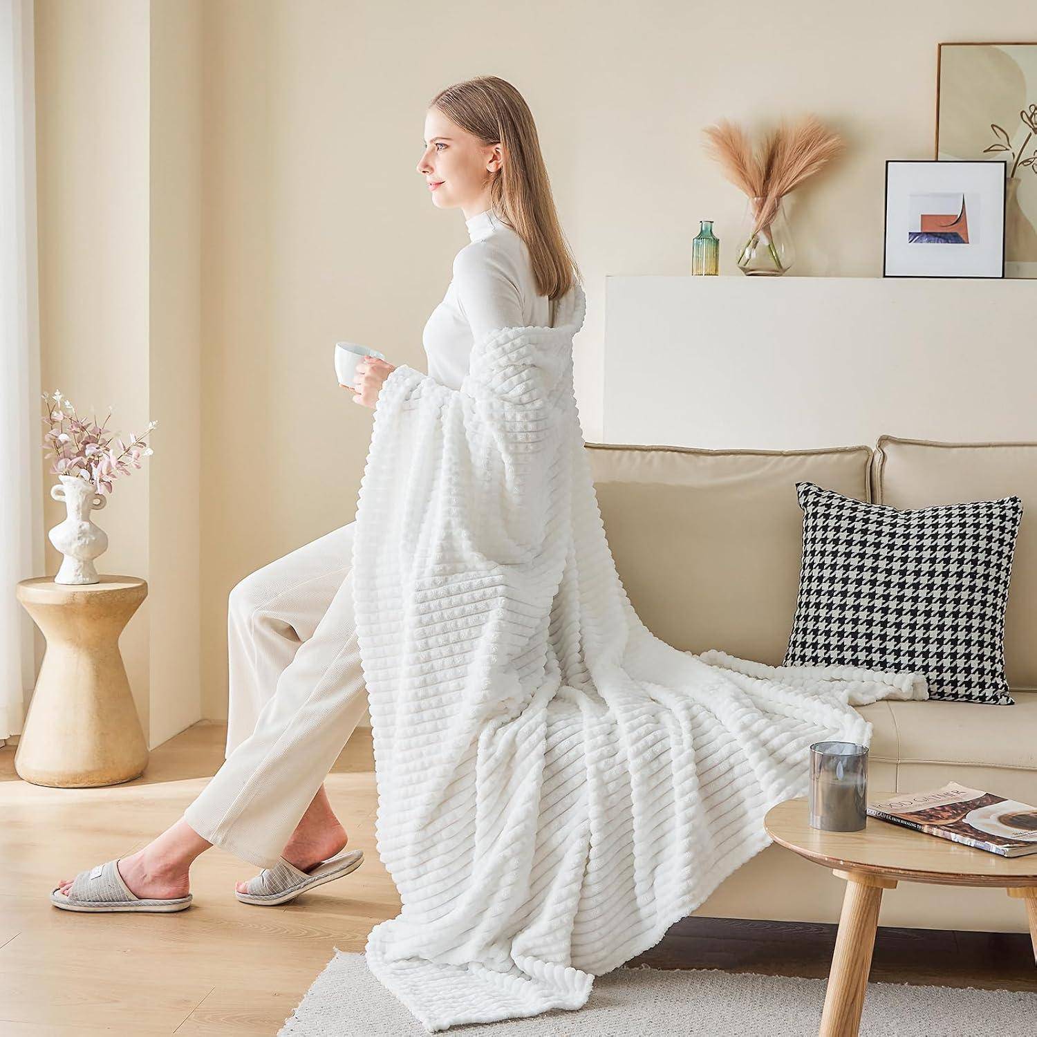 Eine Frau, die in eine gemütliche weiße Decke gehüllt ist, steht neben einem Sofa in einem stilvollen Wohnzimmer und hält eine Tasse in der Hand, was ein Gefühl von Komfort und Entspannung hervorruft.