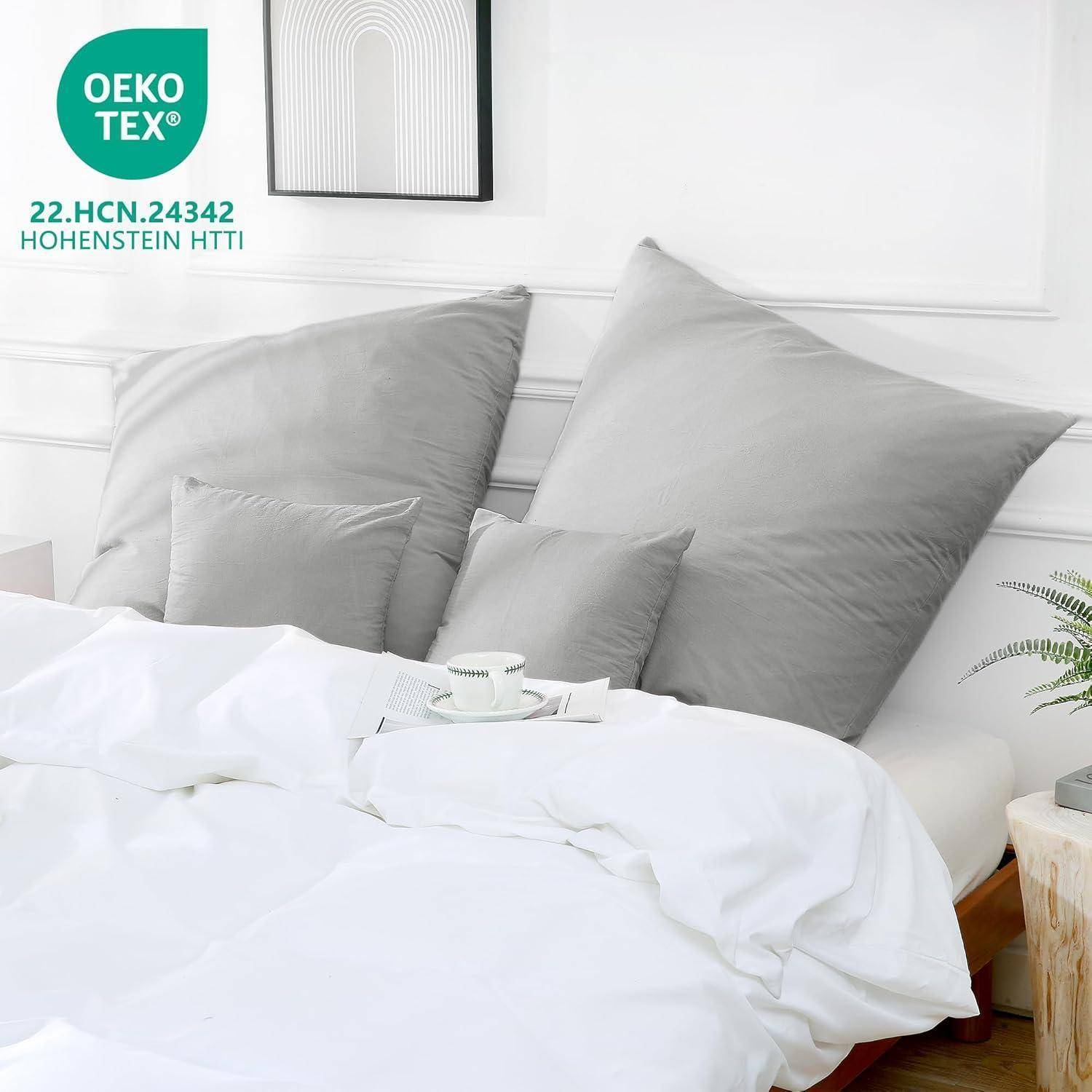 Ein ordentlich gemachtes Bett mit weißen Bettwäsche und großen grauen Kissen an einem weißen Kopfteil, versehen mit der 'OEKO-TEX' Zertifizierung und Code.