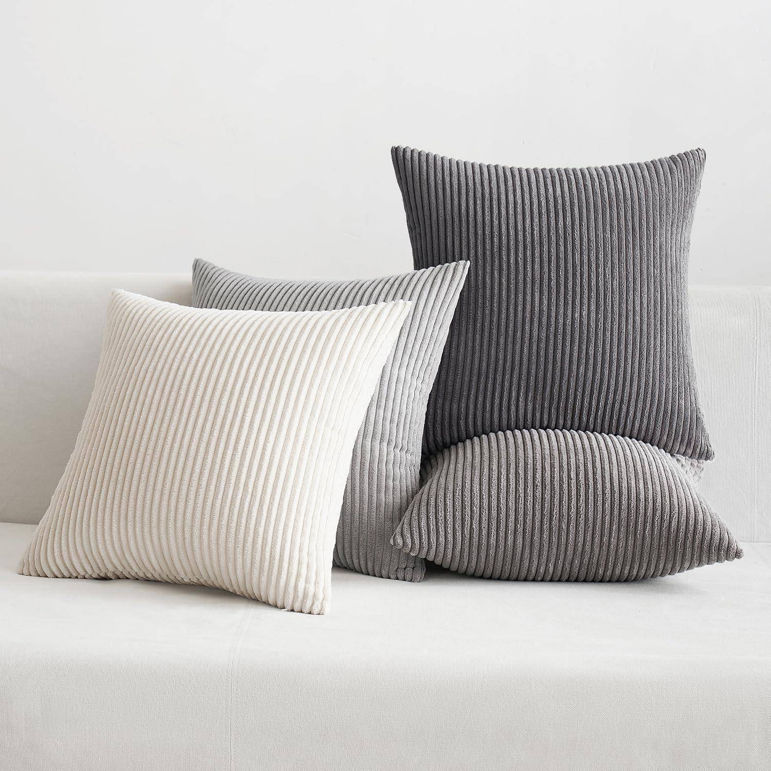 Vier gerippte Kissen in verschiedenen Schattierungen von Grau und Weiß sind auf einem weißen Sofa vor einem schlichten weißen Hintergrund angeordnet.