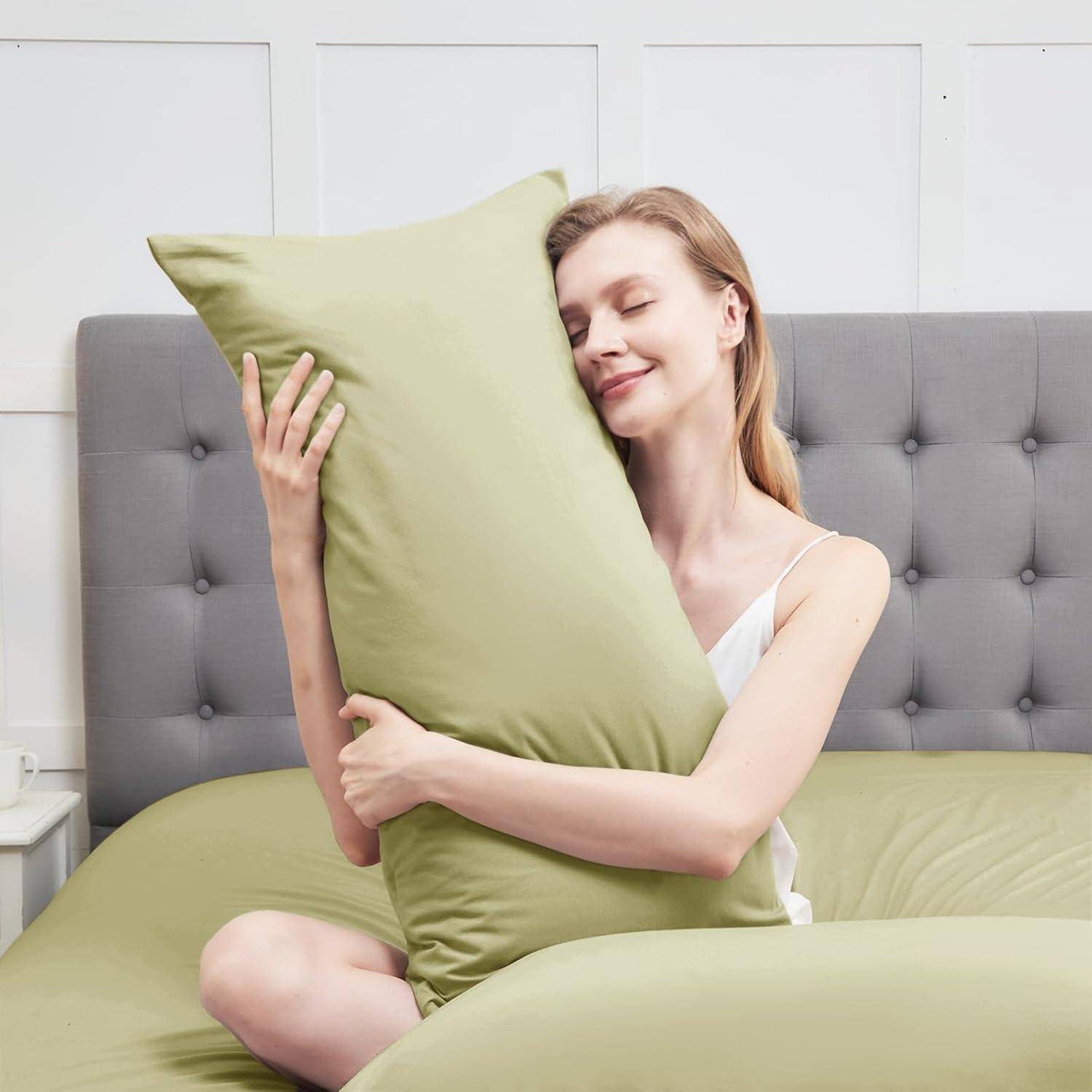 Eine Person sitzt auf einem Bett und umarmt einen grünen Kopfkissen mit einem zufriedenen Ausdruck. Der Raum hat lichtfarbene Wände und Bettwäsche.