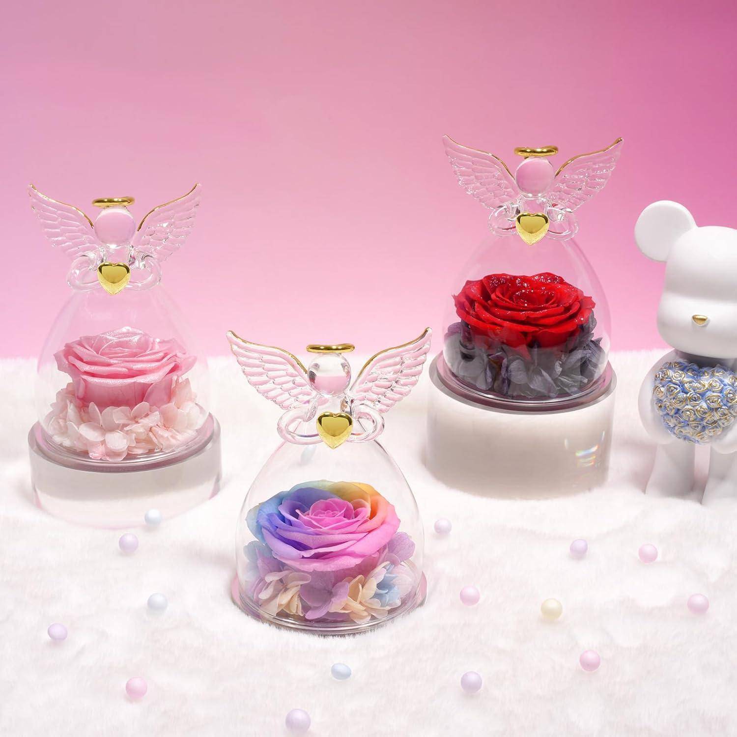 Glasglocken mit Engelflügeln umschließen farbenfroh konservierte Rosen auf einer weichen, weißen Oberfläche. Eine kleine weiße Bärenfigur steht in der Nähe.