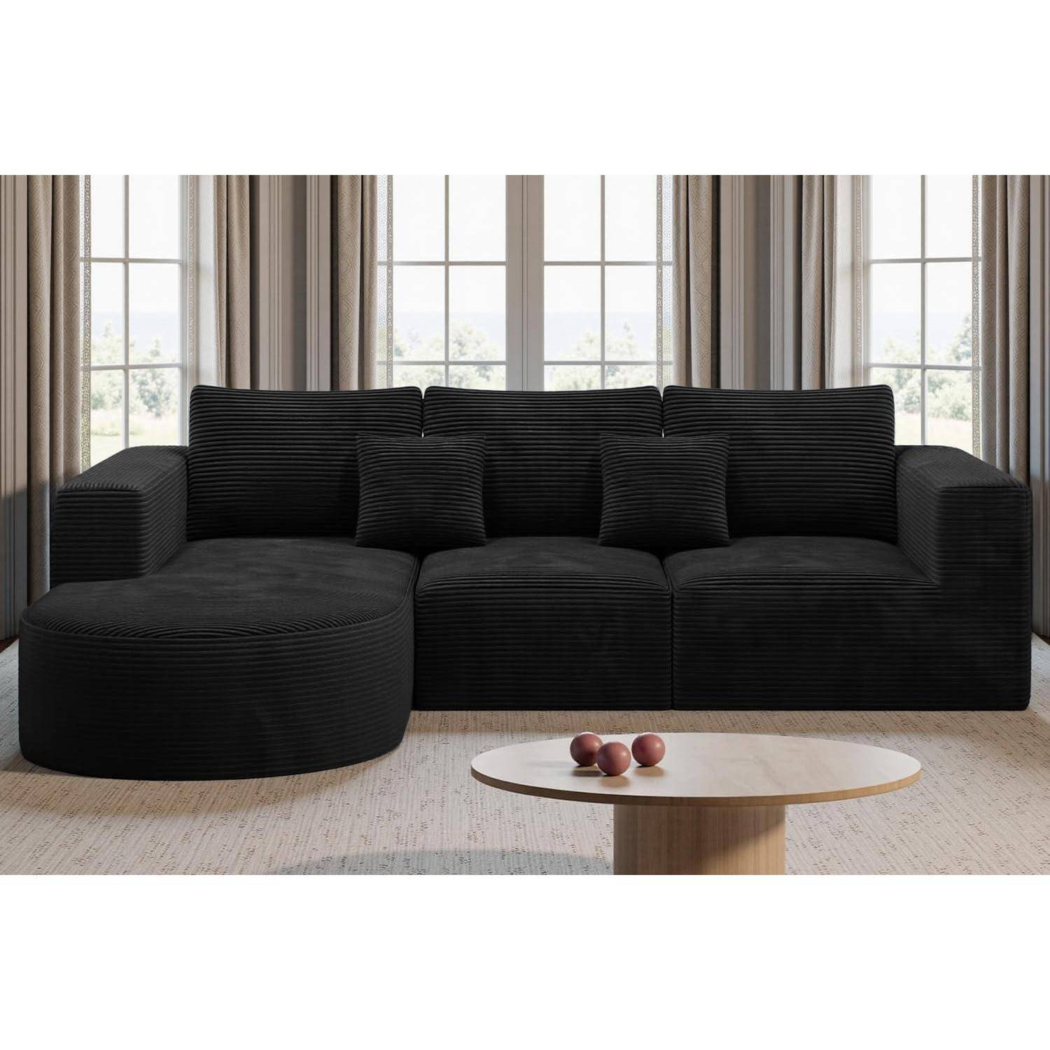 Schwarzes Cordsofa in Wohnlandschaftsform mit Kissen, begleitet von einem runden Holzkaffeetisch, auf dem vier Äpfel liegen, platziert in einem Raum mit hohen Fenstern und Vorhängen.