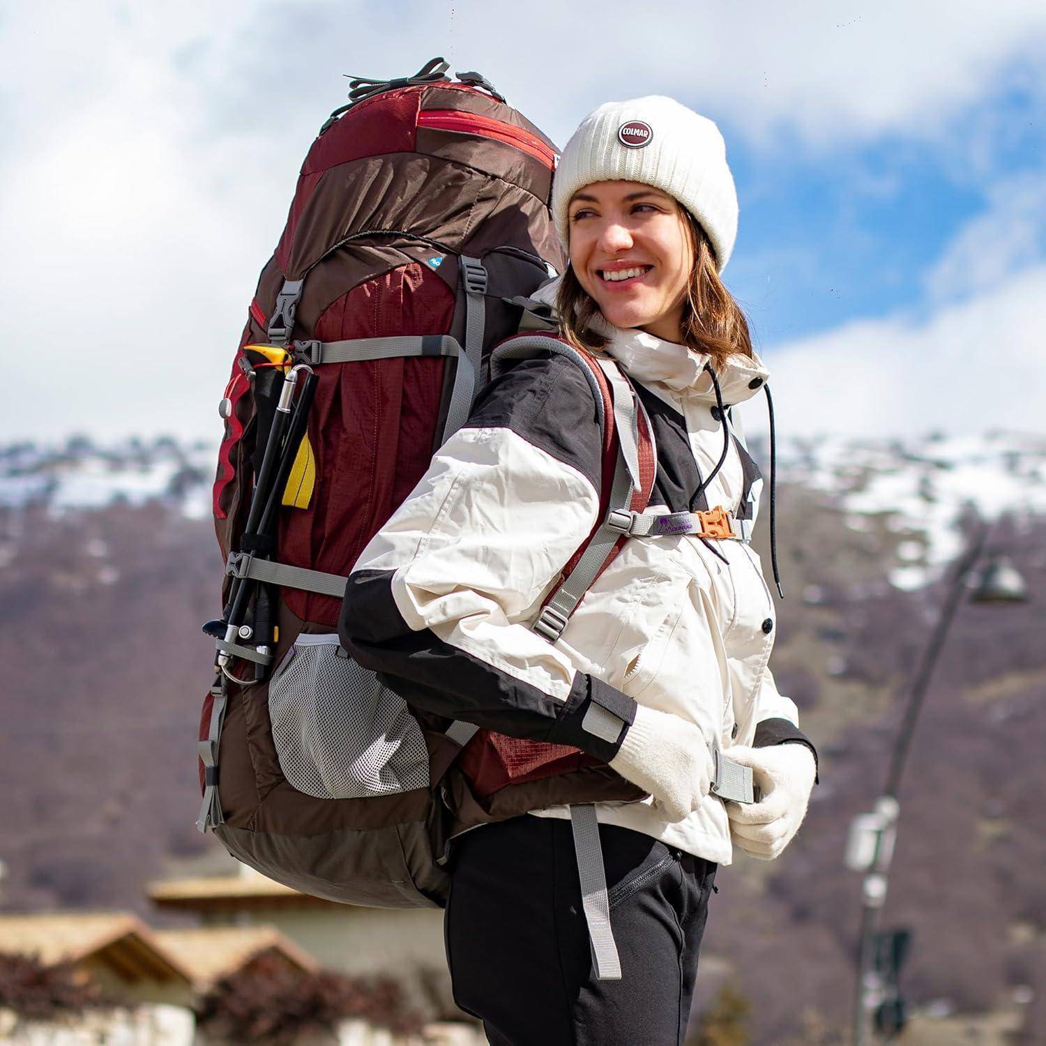 Eine Person mit weißer Jacke und Mütze lächelt beim Wandern mit einem großen Rucksack, mit verschneiten Bergen im Hintergrund.