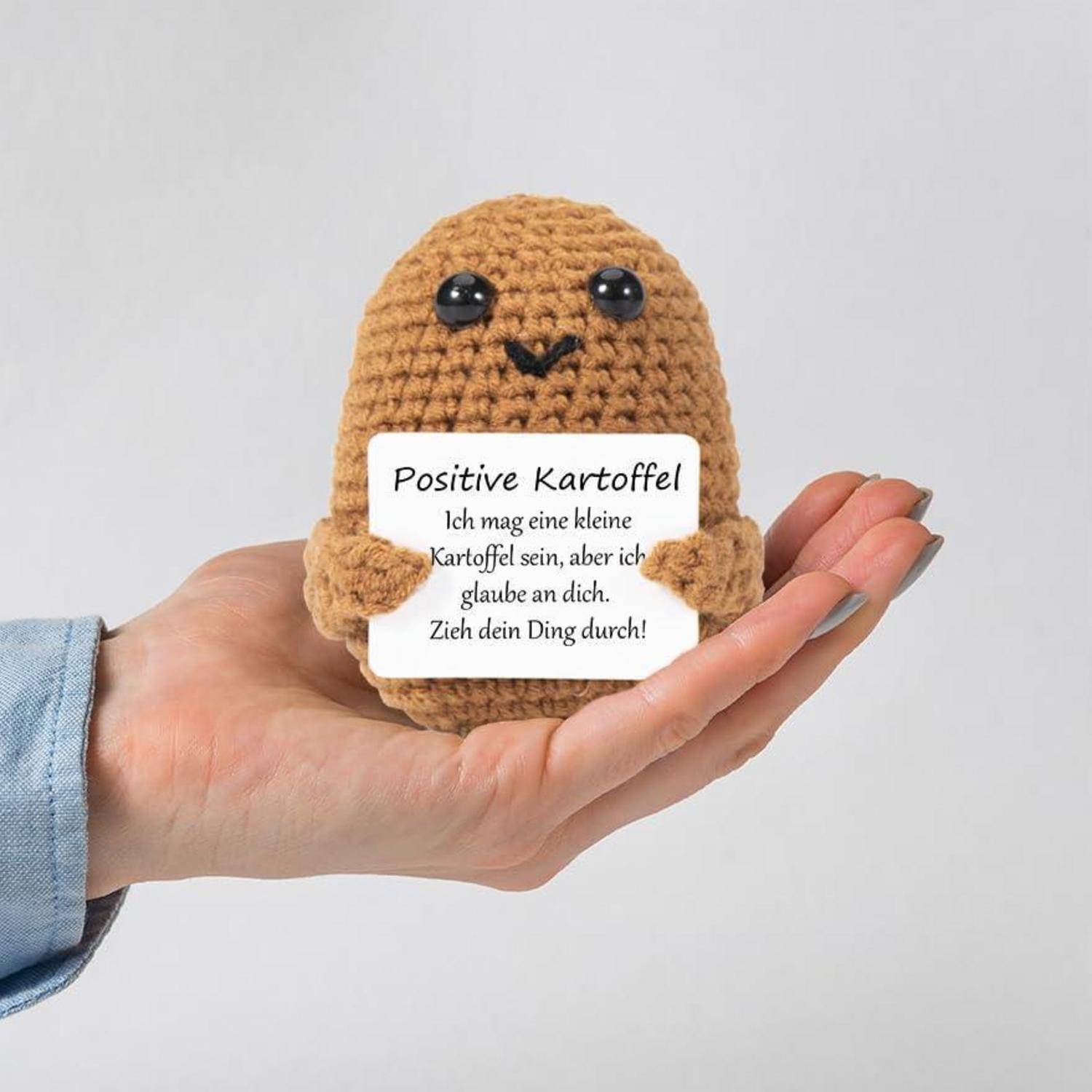 Eine Hand hält ein gestricktes Kartoffelspielzeug mit schwarzen Augen. Das Spielzeug hält ein Schild mit der Aufschrift: 