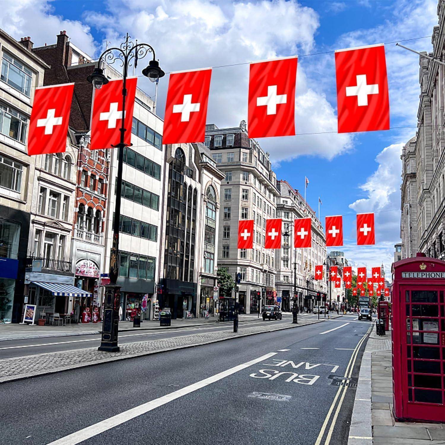 Eine Stadtstraße mit Schweizer Flaggen, die darüber hängen; historische Gebäude säumen die Seiten, eine rote britische Telefonzelle rechts, unter einem strahlend blauen Himmel.