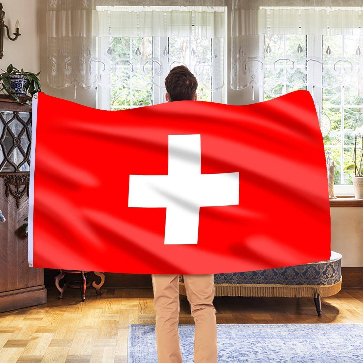 Eine Person steht drinnen und hält eine große Schweizer Flagge mit einem weißen Kreuz auf rotem Hintergrund, die zu einem sonnenbeschienenen Fenster blickt.