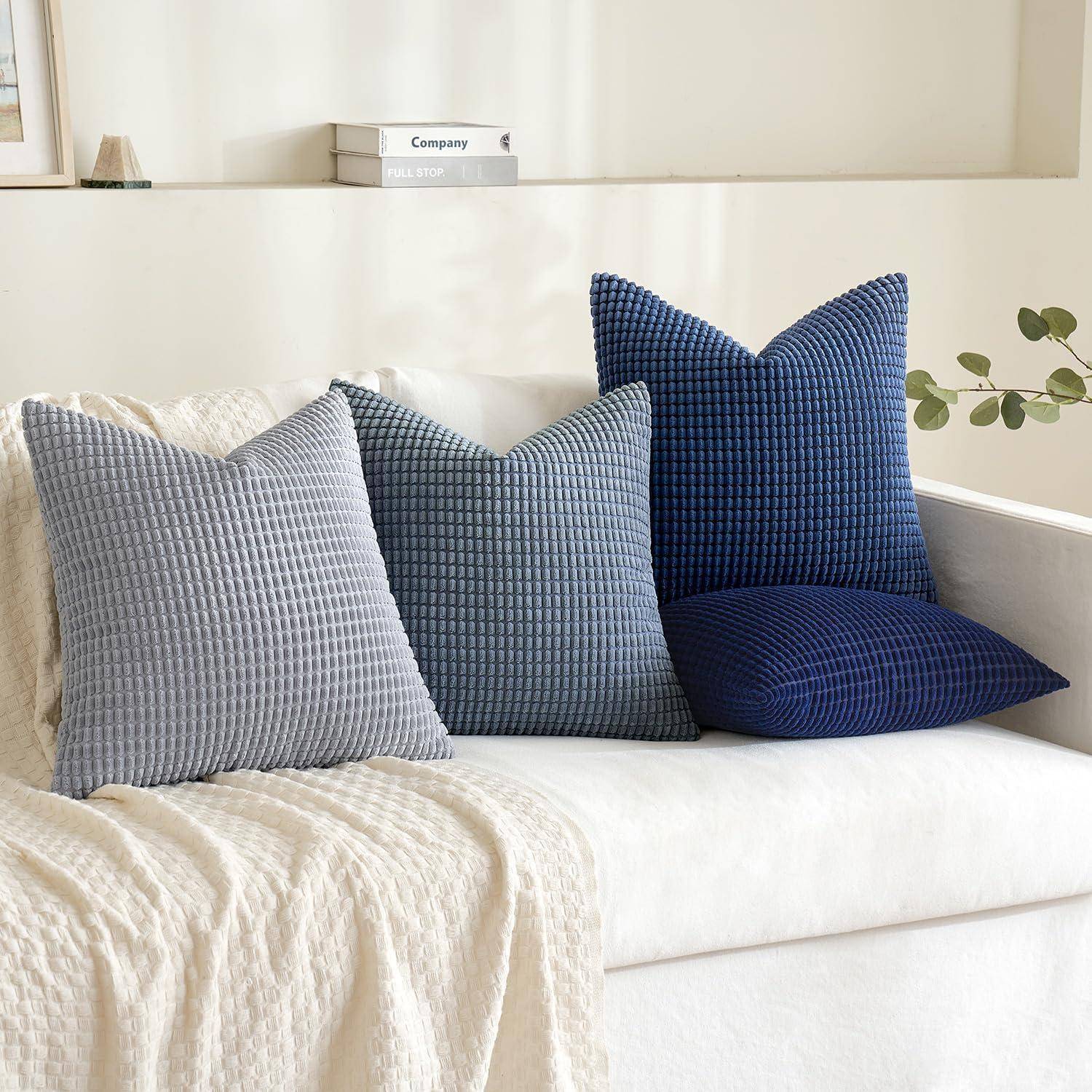 Zierkissen in strukturierten blauen und grauen Stoffen sind auf einem weißen Sofa angeordnet, wobei im Hintergrund eine Pflanze und ein kleines Regal zu sehen sind.