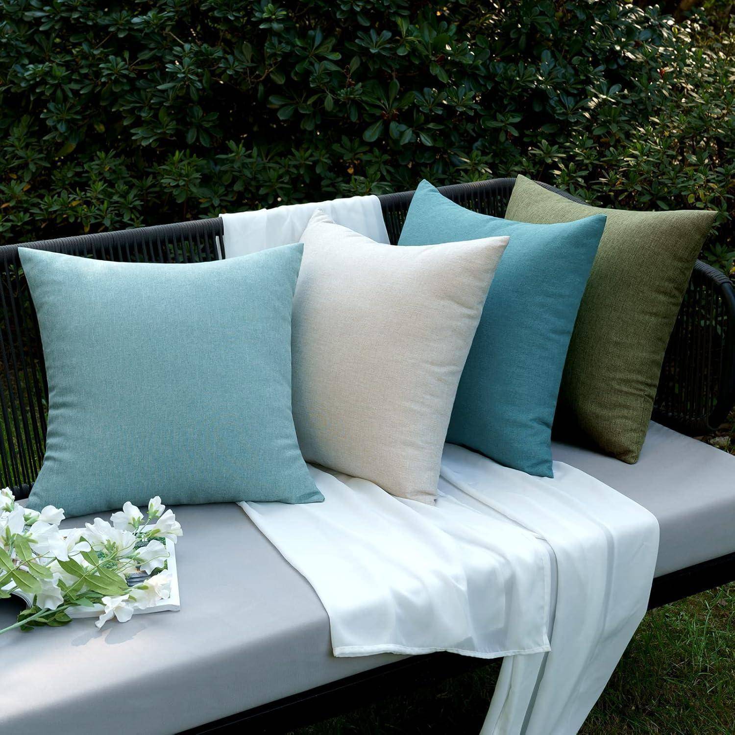 Ein Set aus vier dekorativen Kissen in Blau, Creme, Petrol und Grün auf einer dunklen Bank mit einem weißen Tuch und künstlichem Grün für eine gemütliche Atmosphäre.
