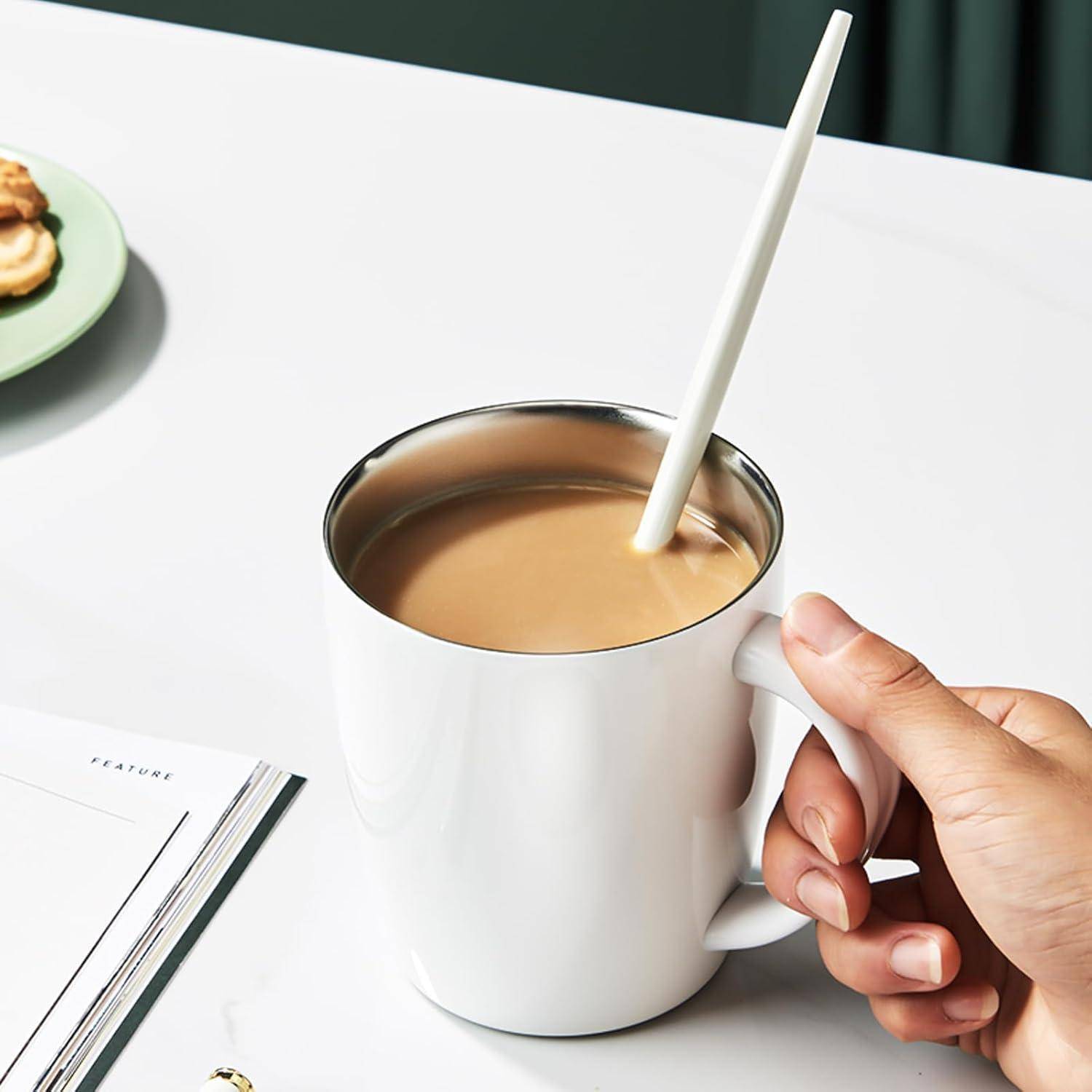 Eine Hand, die eine weiße Tasse gefüllt mit Kaffee hält, ein Strohhalm darin. Ein Notizbuch und ein Keks auf einem Teller liegen daneben auf einem Tisch.