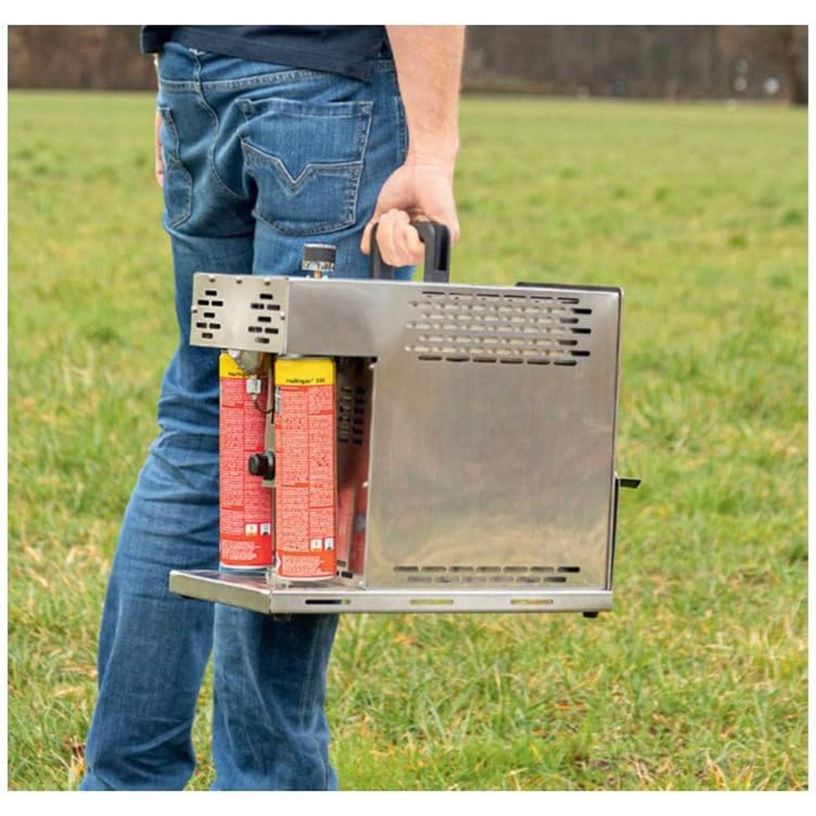 Eine Person, die einen tragbaren Campingkocher mit zwei Brennstoffkanistern über ein Grasfeld trägt.