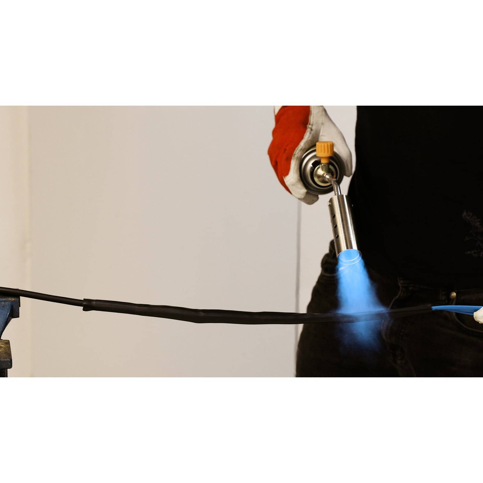 Eine Person benutzt einen Schweißbrenner, um eine Metallstange zu erhitzen. Der Schweißbrenner emittiert eine blaue Flamme. Die Person trägt Schutzhandschuhe.