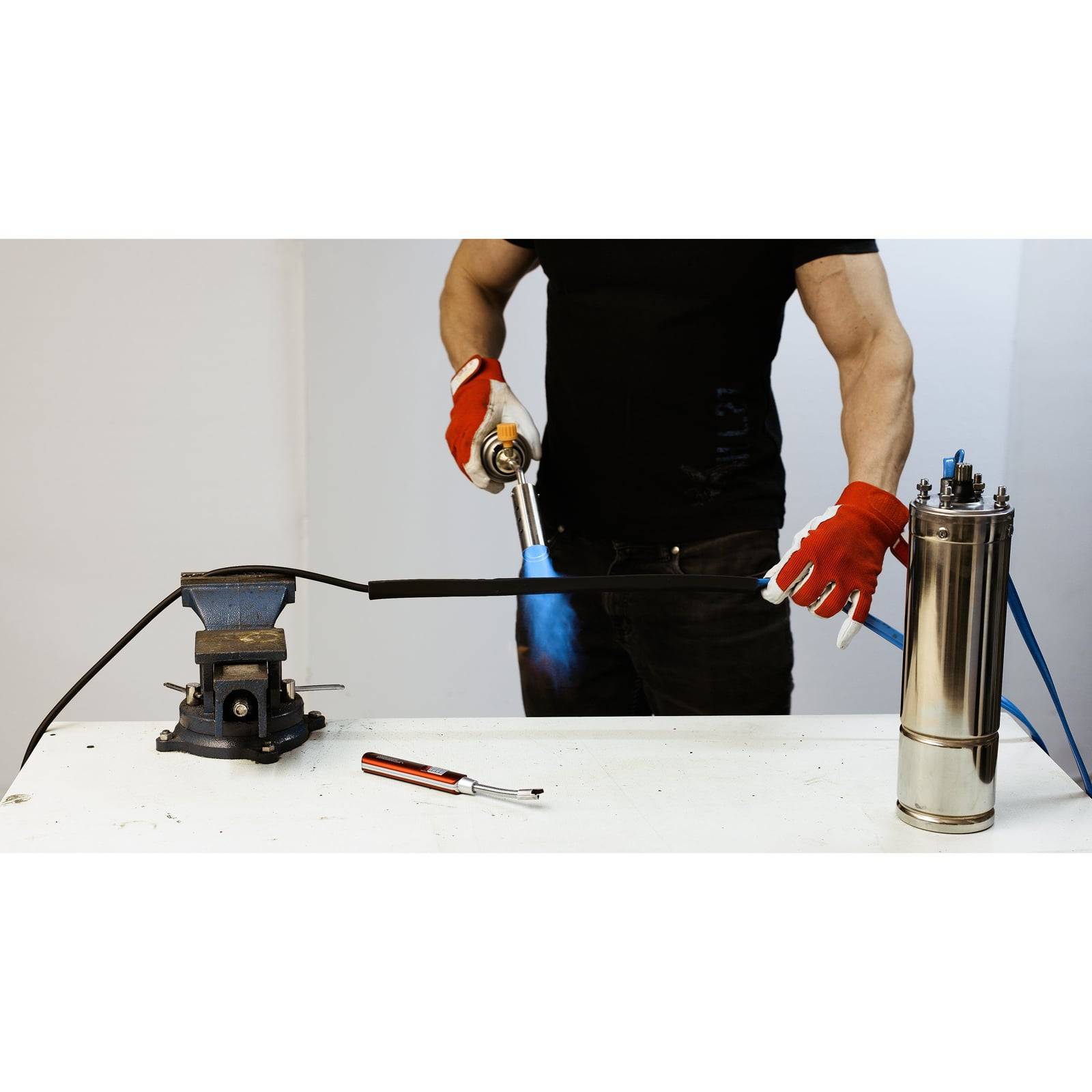 Eine Person mit roten Handschuhen, die in einer Werkstatt einen Brenner verwendet, um ein Kabel zu erhitzen. Ein Schraubstock und Werkzeuge sind auf dem Tisch sichtbar.