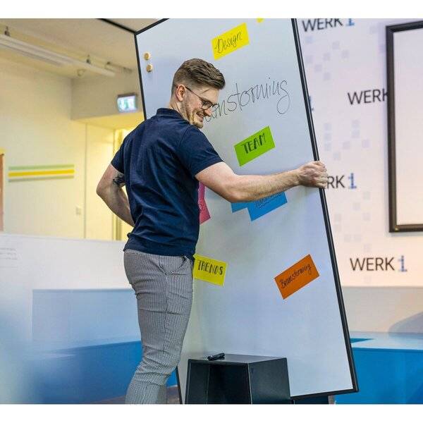 Ein Mann in einer Werkstatt bewegt ein Whiteboard mit Klebezetteln, auf denen Begriffe wie „Design