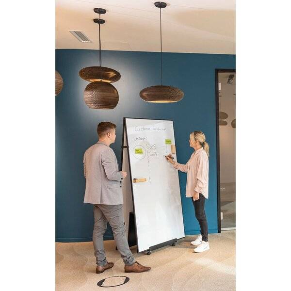 Zwei Kollegen besprechen Notizen auf einem Whiteboard in einem modernen Büroflur, wobei die Frau auf die Tafel zeigt und der Mann zuhört.