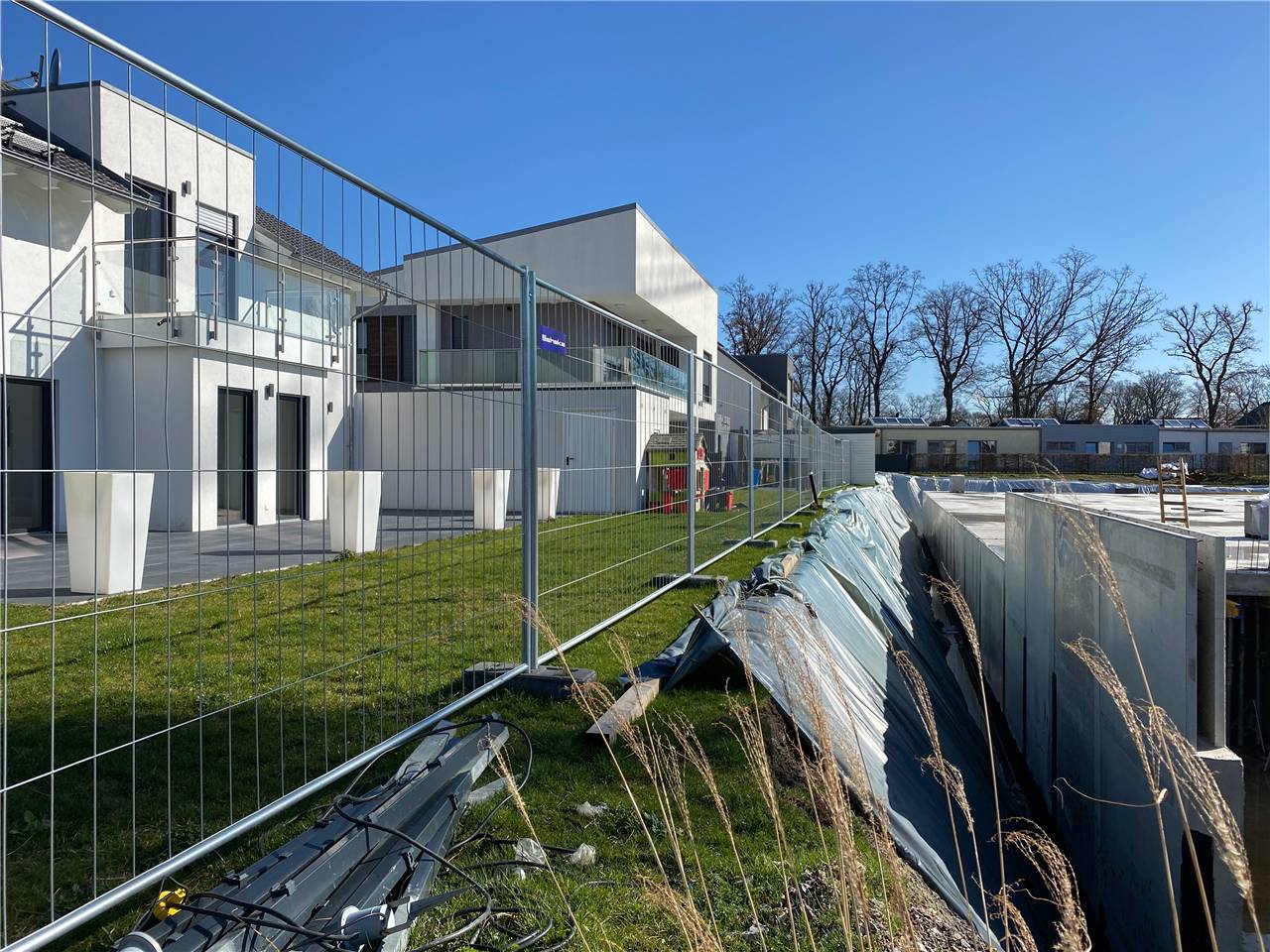 Baustelle mit modernem Gebäude, abgesperrtem Bereich und mit Planen abgedeckten Materialien. Kahle Bäume im Hintergrund unter klarem Himmel.