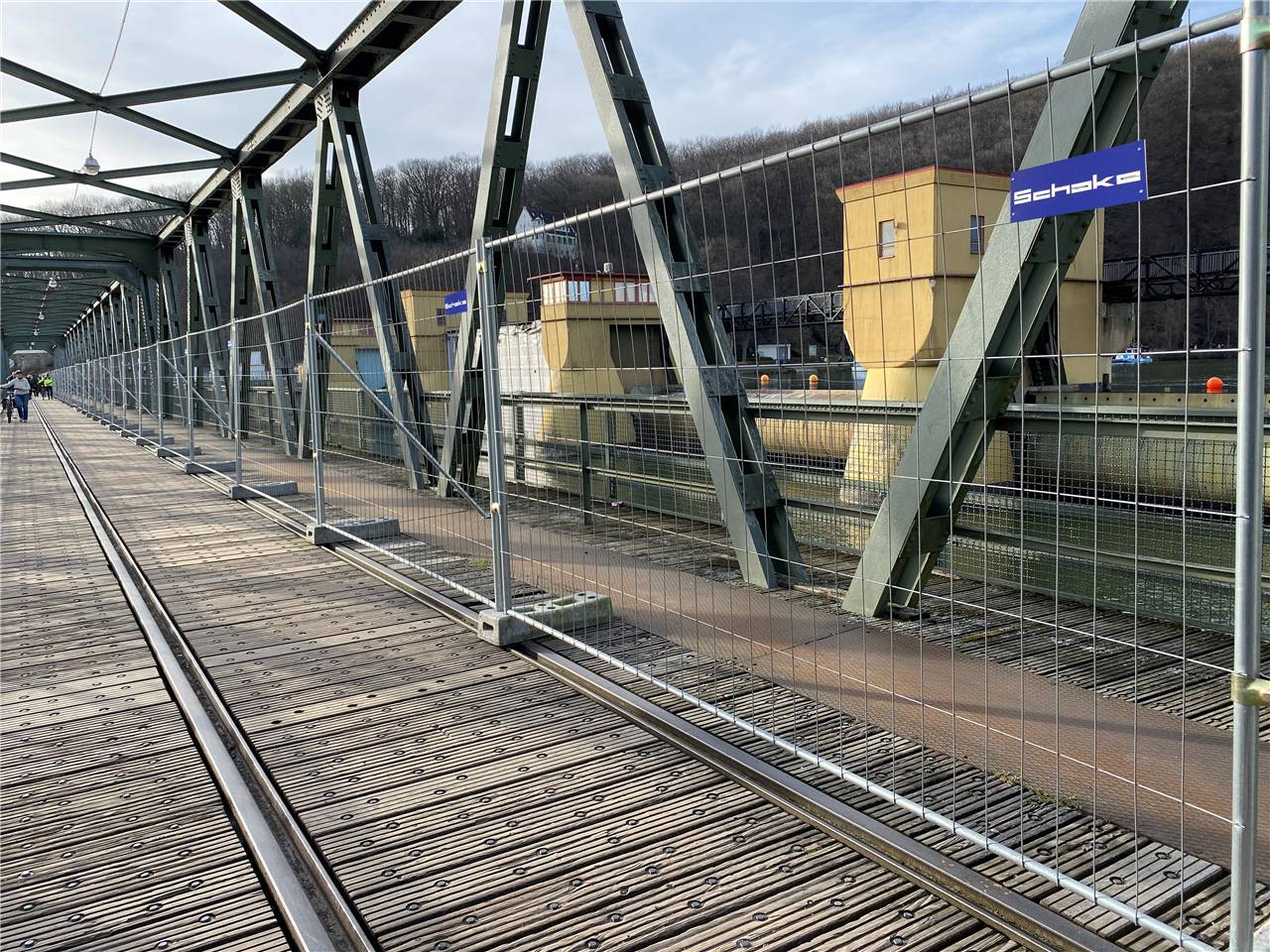 Eine Fußgängerbrücke mit Metallstützen und Holzbrettern überspannt eine Wasserstraße. Gebäude und Hügel sind im Hintergrund unter einem bewölkten Himmel zu sehen.