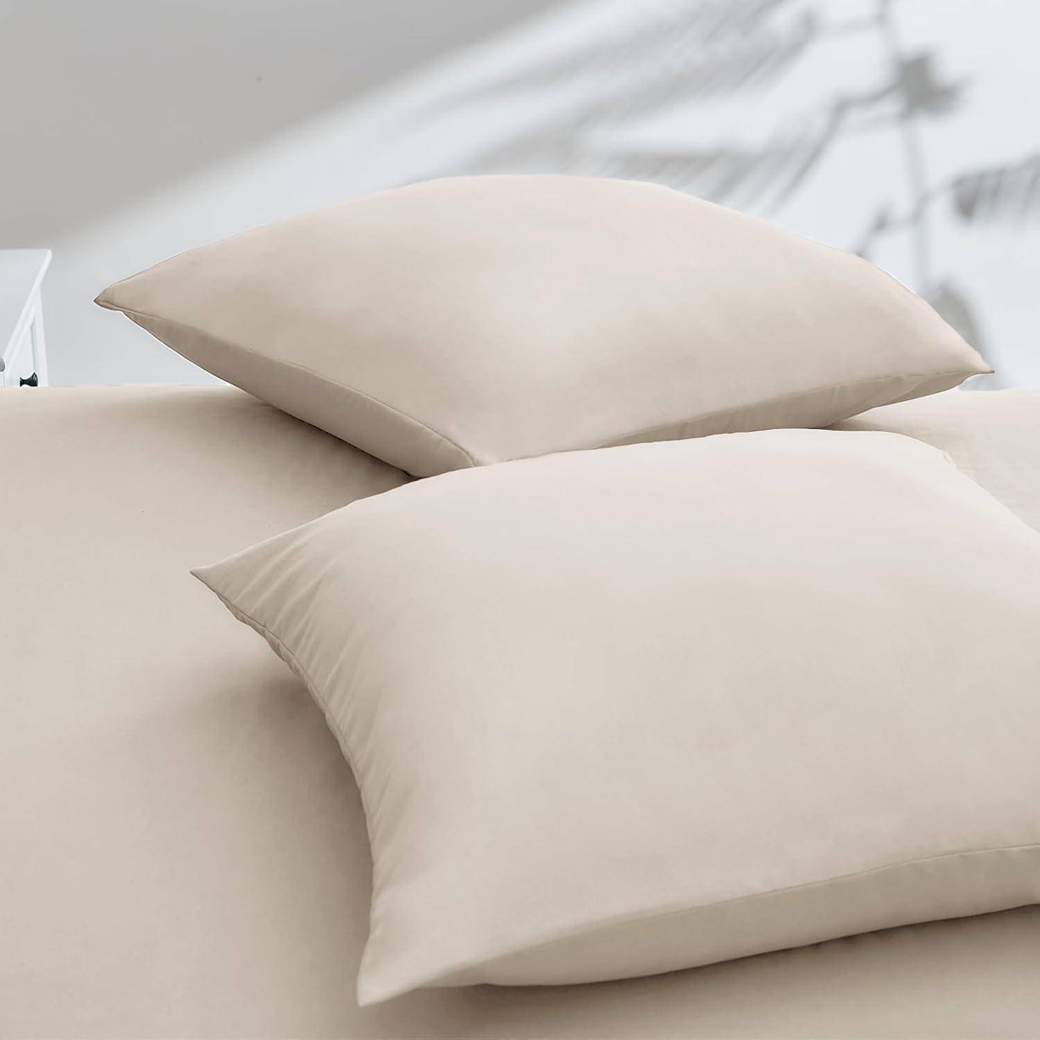 Zwei beige Kissen auf einem Bett mit einem weichen, weißen Überwurf, unter natürlichem Licht. Schatten von Pflanzenbättern erscheinen an der Wand dahinter.