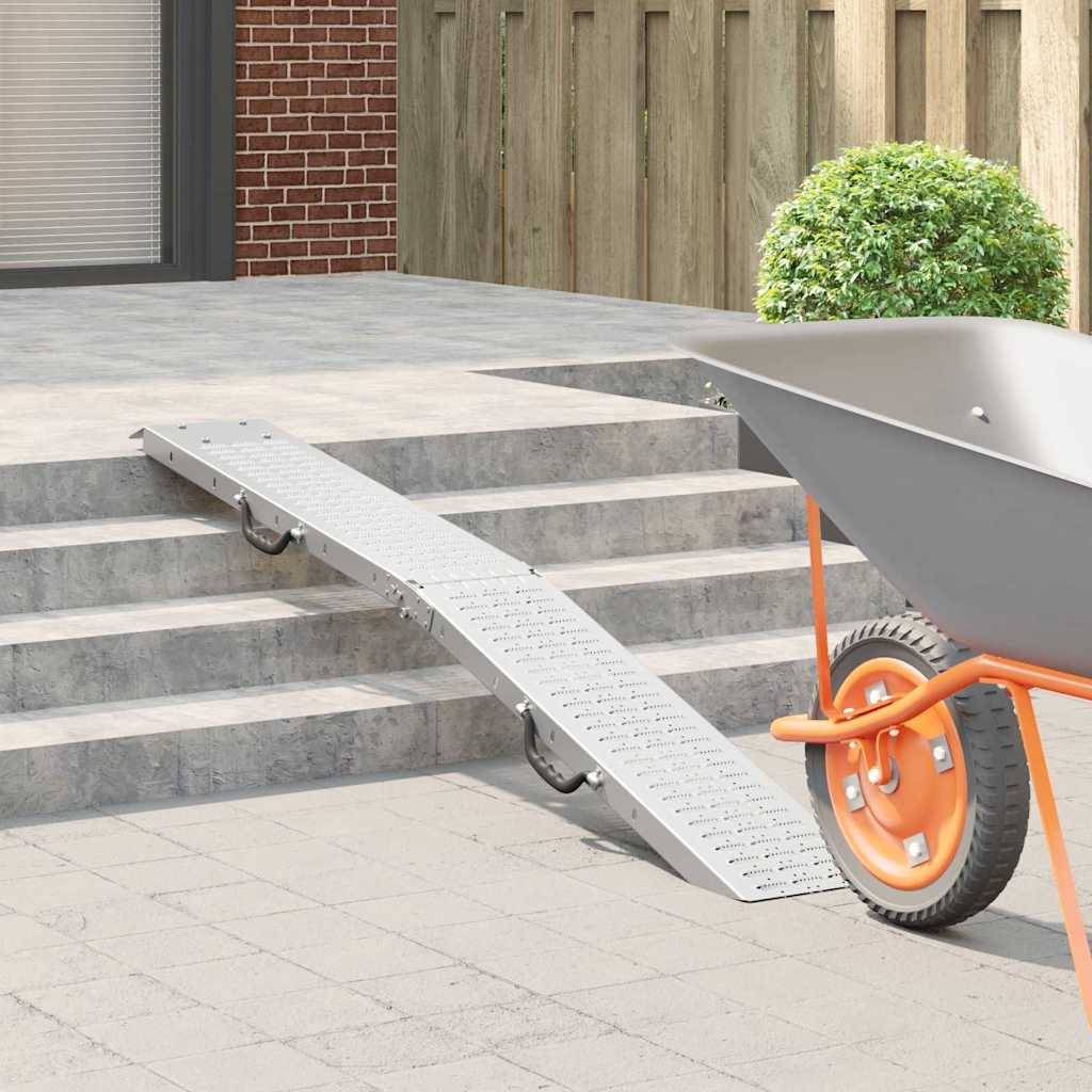 Eine Metallrampe führt zu einer kleinen Treppe vor dem Gebäudeeingang und erleichtert so die Barrierefreiheit. Eine Schubkarre ist in der Nähe platziert.