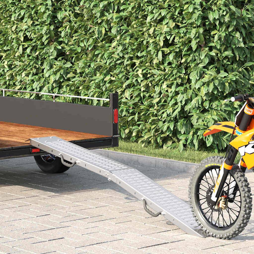 Ein Motorrad ist am Fuß einer Metallrampe geparkt, die zu einem Anhänger führt, vor einer Kulisse von Grünpflanzen auf einer gepflasterten Oberfläche.