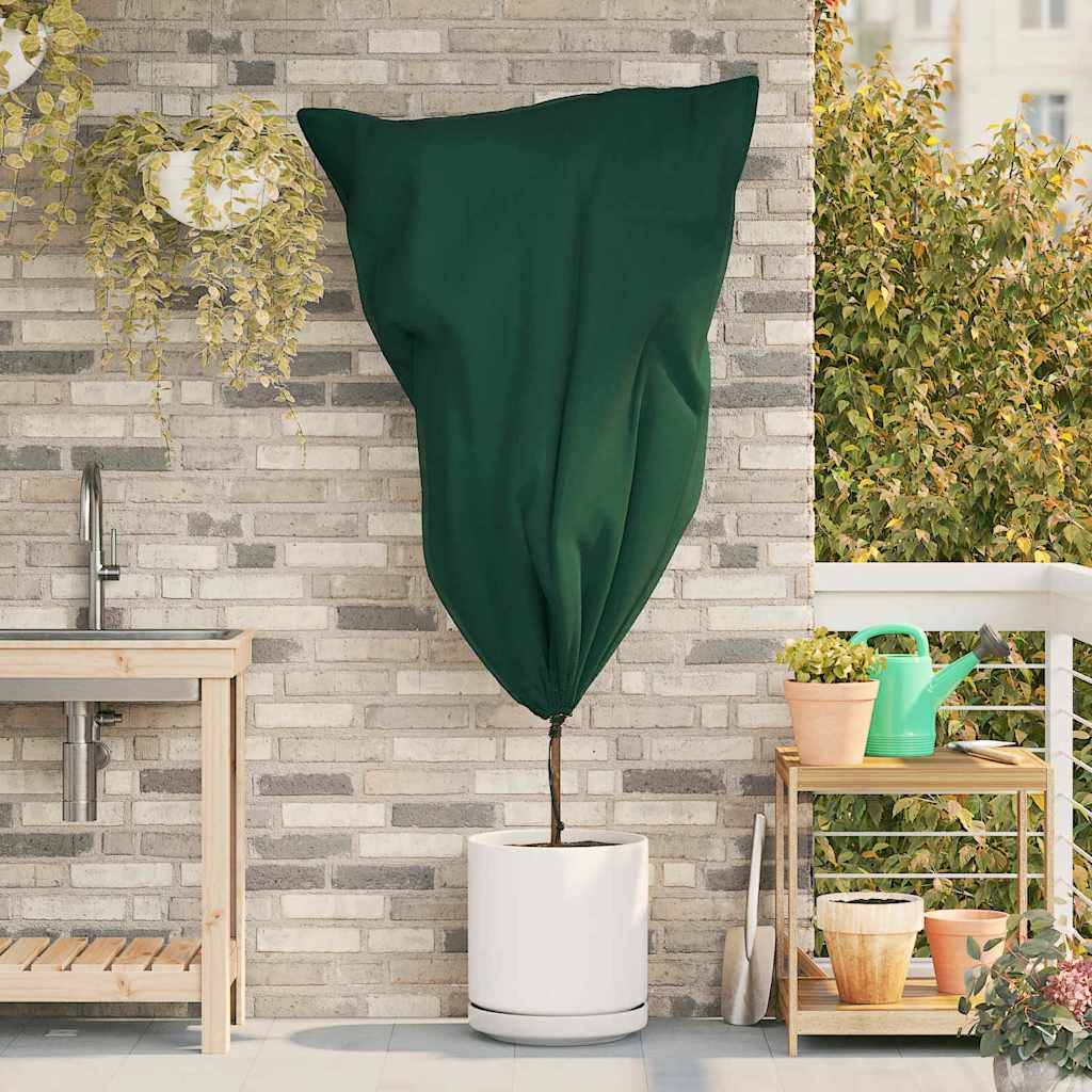 Eine hohe Pflanze mit einer dunkelgrünen Stoffabdeckung in einem weißen Topf vor einer Ziegelwand mit einem Wasserhahn. Gartenwerkzeuge und eine Gießkanne in der Nähe.