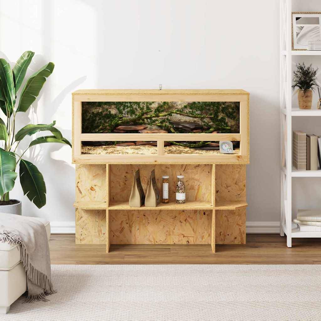 Ein hölzernes Terrarium steht auf einem Ständer in einem gut beleuchteten Wohnzimmer, mit einer Wüstenlandschaft im Inneren. Links befindet sich eine Pflanze, rechts sind Regale.