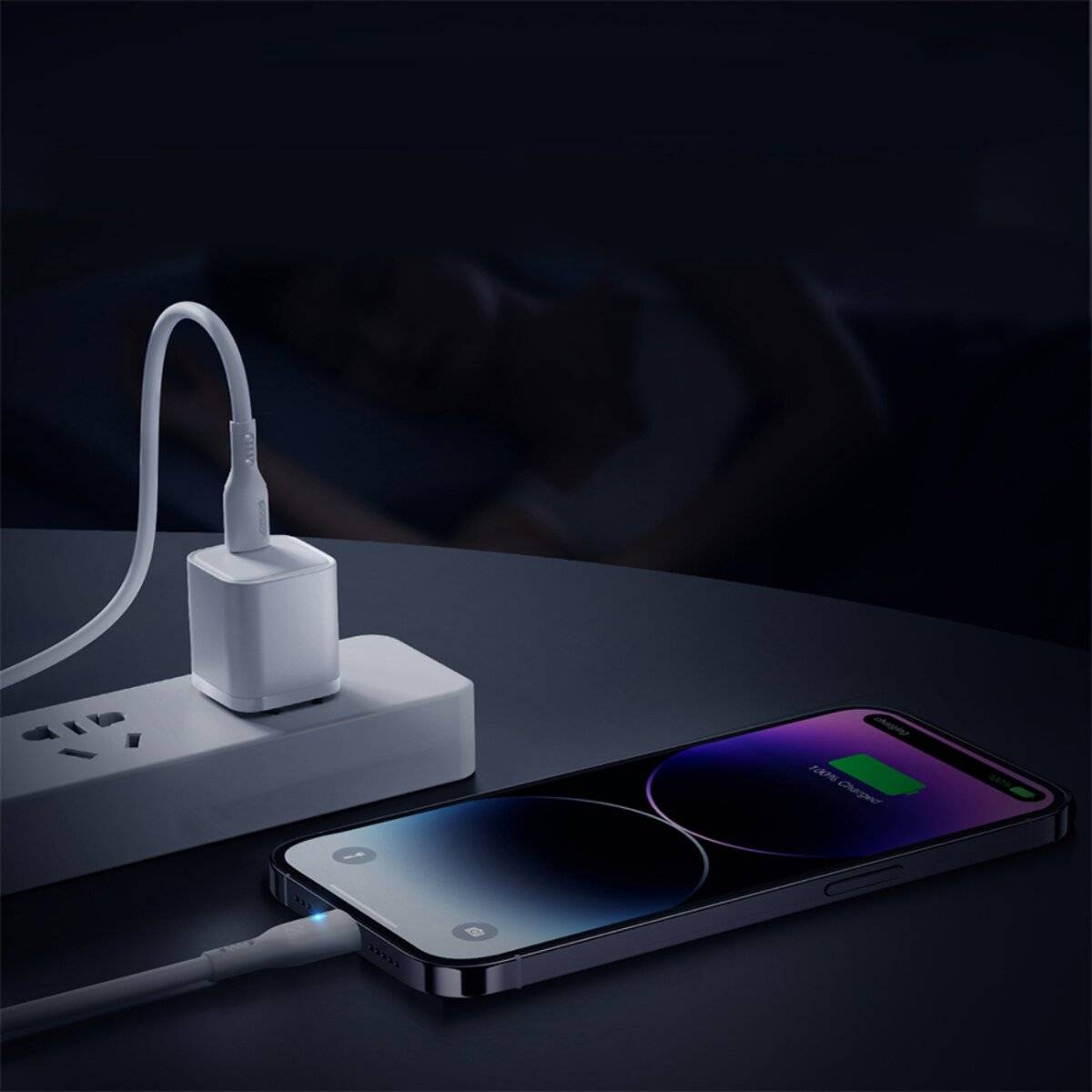 Ein Smartphone lädt nachts auf einem Schreibtisch, während im Hintergrund eine Person schläft. Der Bildschirm des Telefons zeigt ein Ladensymbol.