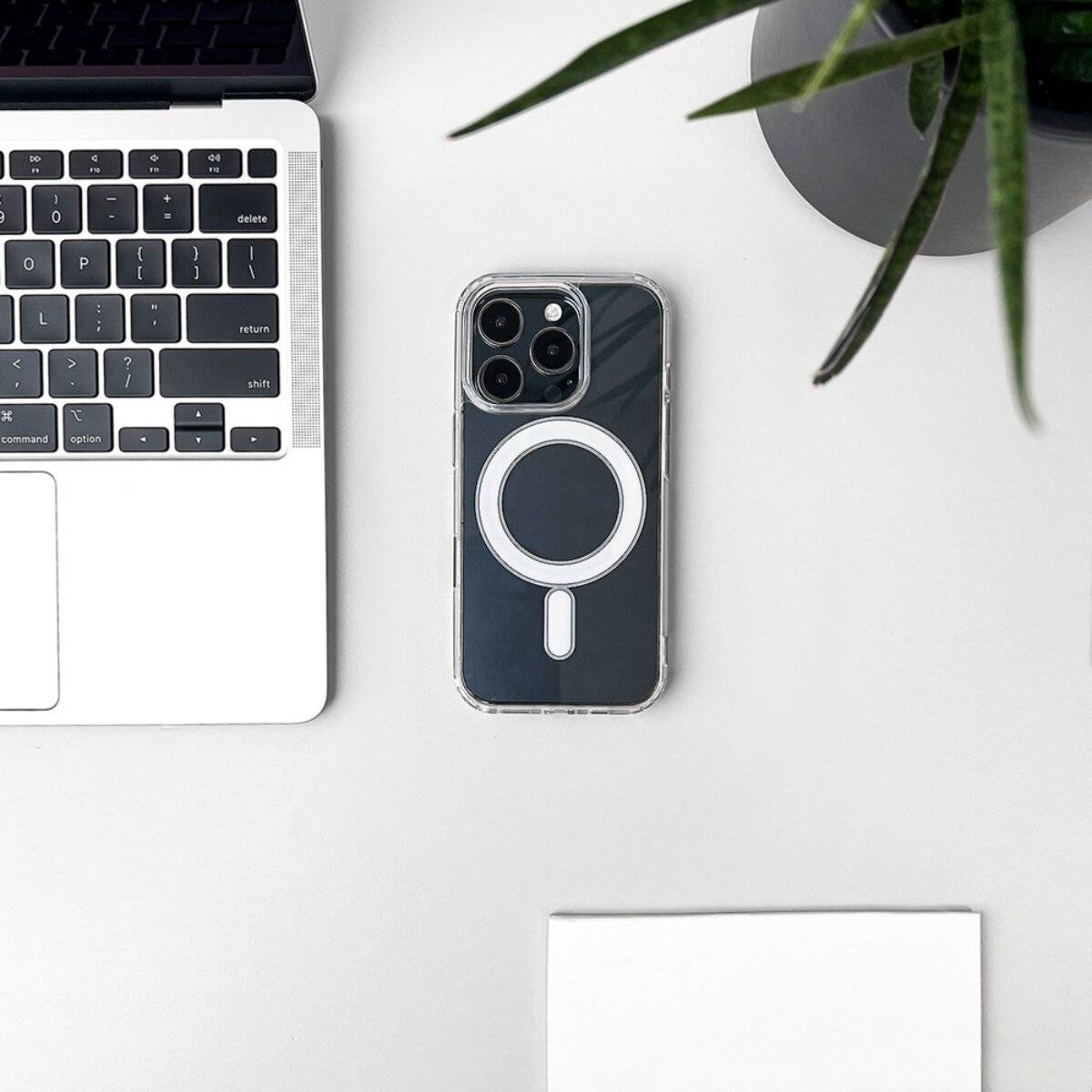 Eine Laptoptastatur, ein Telefon mit einem MagSafe-Etui, eine Topfpflanze und ein weißes Notizbuch sind ordentlich auf einem weißen Schreibtisch angeordnet.