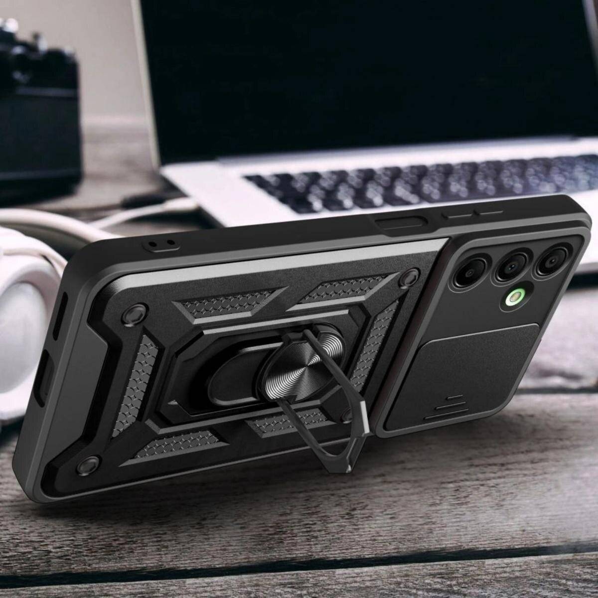 Ein Smartphone mit einem robusten schwarzen Gehäuse, das einen integrierten Ringständer besitzt, ist auf einem Holztisch neben einem Laptop aufgestellt.