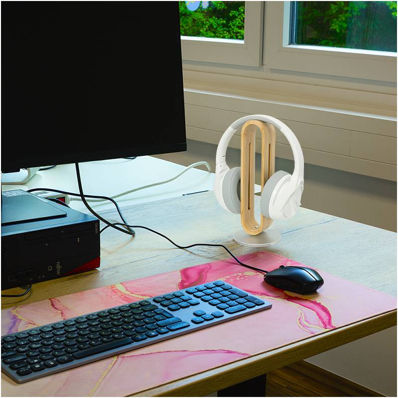 Computerarbeitsplatz mit einem Monitor, Tastatur, Maus auf einer rosa Matte und einem weißen Kopfhörerständer neben einem Fenster mit Blick auf Grünflächen.