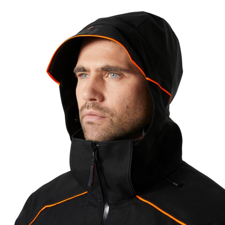 Eine Person mit einer schwarzen Kapuzenjacke mit orangefarbenen Akzenten, die konzentriert in die Ferne blickt, vor einem schlichten Hintergrund.