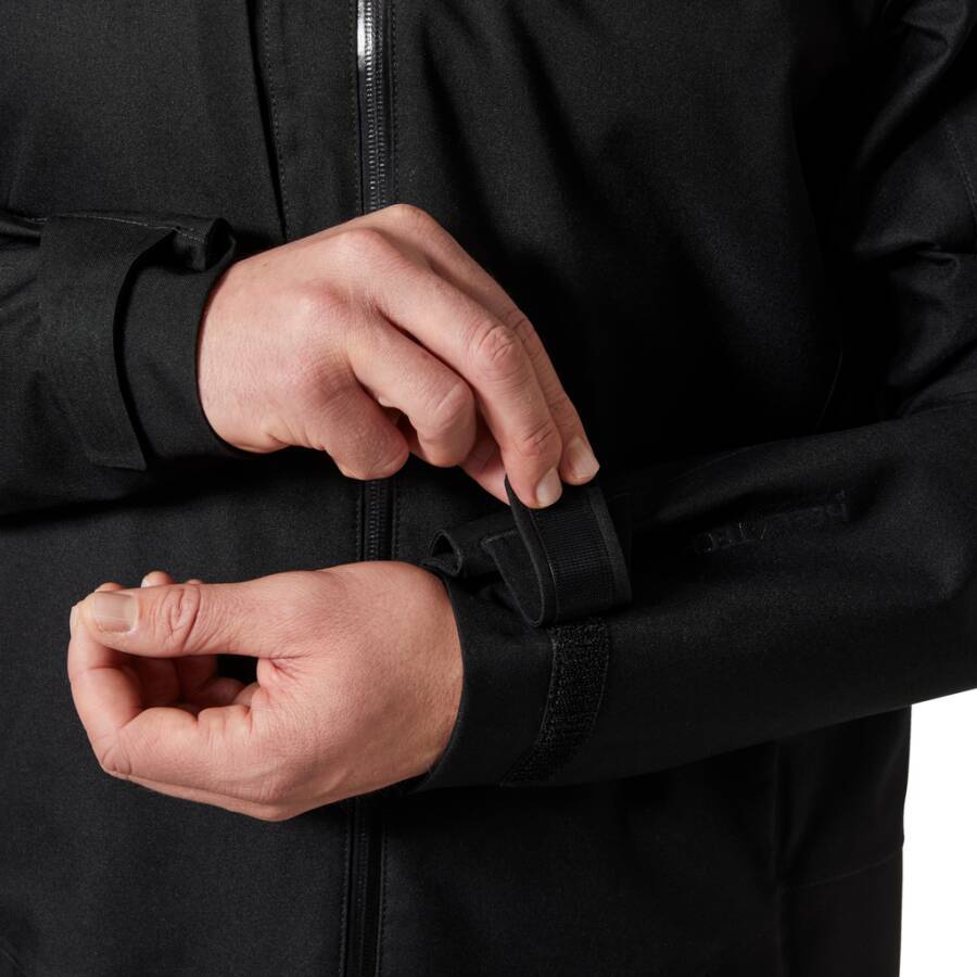 Hände, die den Manschettengurt an einer schwarzen Jacke einstellen, mit Fokus auf das funktionale Detail des Befestigens der Passform am Handgelenk.