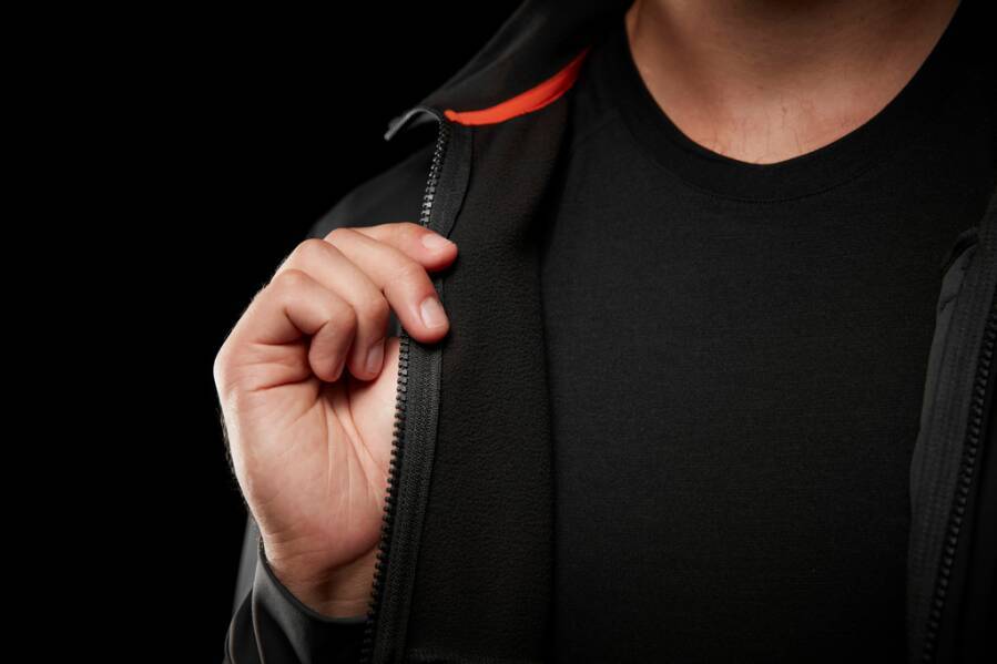 Eine Person zieht einen Reißverschluss einer Jacke teilweise zu und konzentriert sich dabei auf die Hand und den Reißverschluss, wobei ein schwarzes Shirt darunter sichtbar ist.