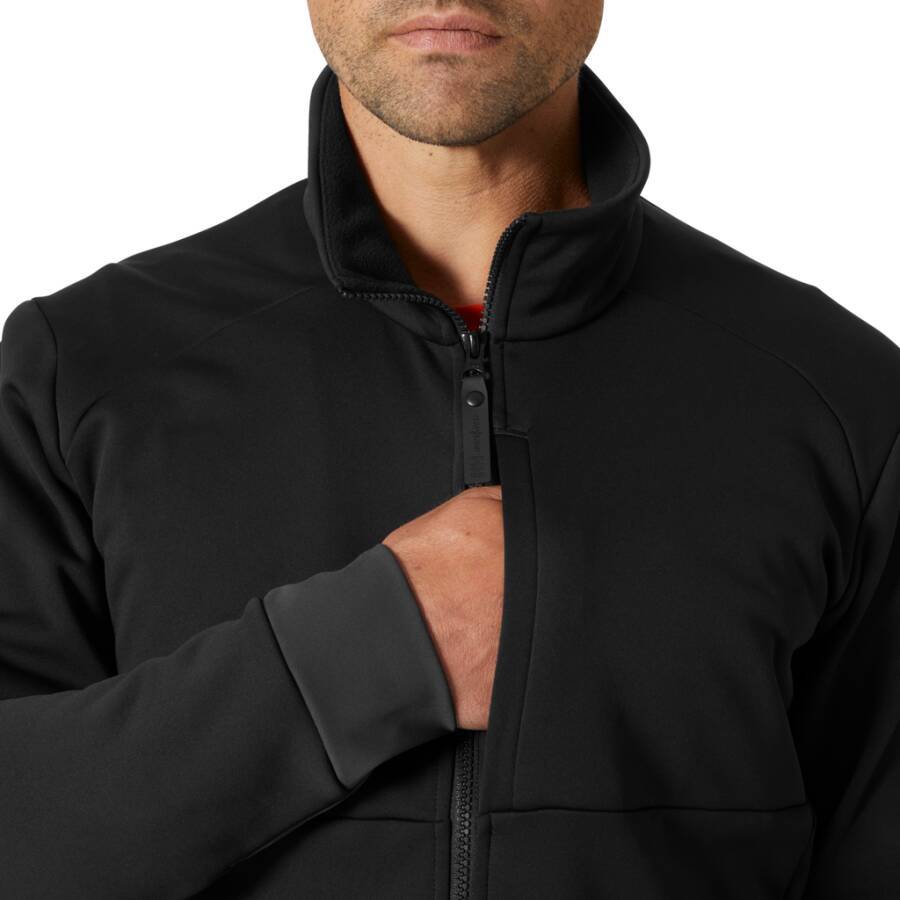 Eine Person, die eine schwarze Reißverschlussjacke trägt, teilweise geöffnet, sodass ein Hauch eines roten Hemdes darunter zu sehen ist, vor einem einfarbigen Hintergrund.