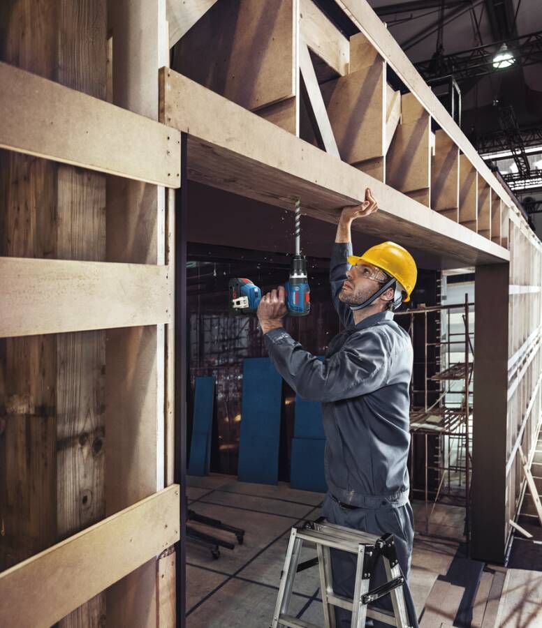 Ein Bauarbeiter mit Schutzhelm bedient eine Bohrmaschine an einem Holzgerüst, steht auf einer Leiter, wobei Baugerüste im Hintergrund sichtbar sind.