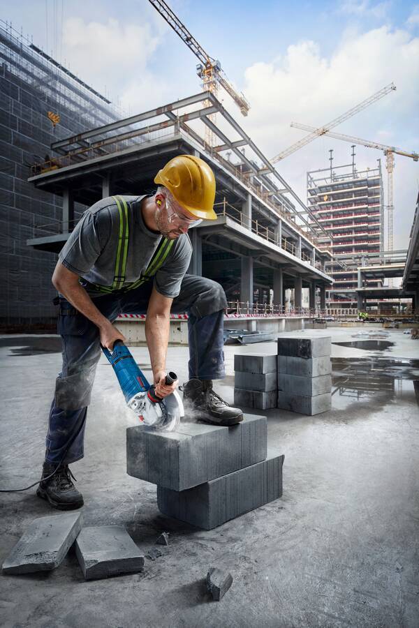 Ein Bauarbeiter in Schutzausrüstung verwendet eine Kreissäge auf Betonblöcken an einer aktiven Baustelle mit Kränen und Gebäuden im Hintergrund.