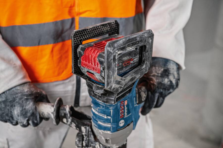 Ein Bauarbeiter in einer orangefarbenen Sicherheitsweste und Handschuhen bedient eine Bosch-Bohrmaschine mit einem rot-schwarzen Aufsatz im Innenbereich.