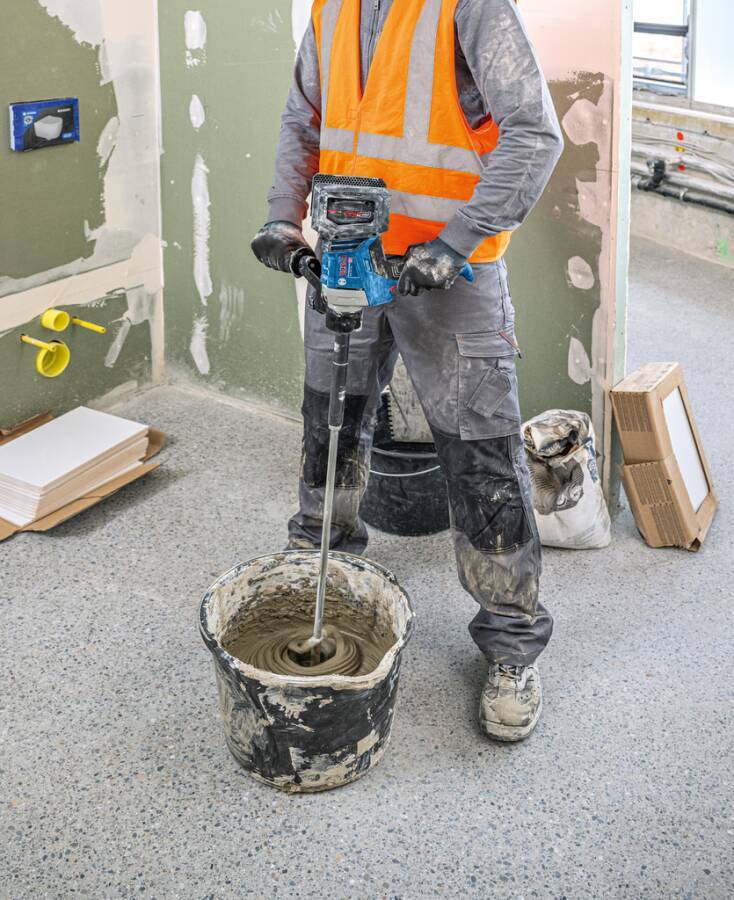 Ein Bauarbeiter in einer orangefarbenen Warnweste benutzt ein Elektrowerkzeug, um eine Substanz in einem Eimer auf einem Betonboden neben einer Wand zu vermischen.