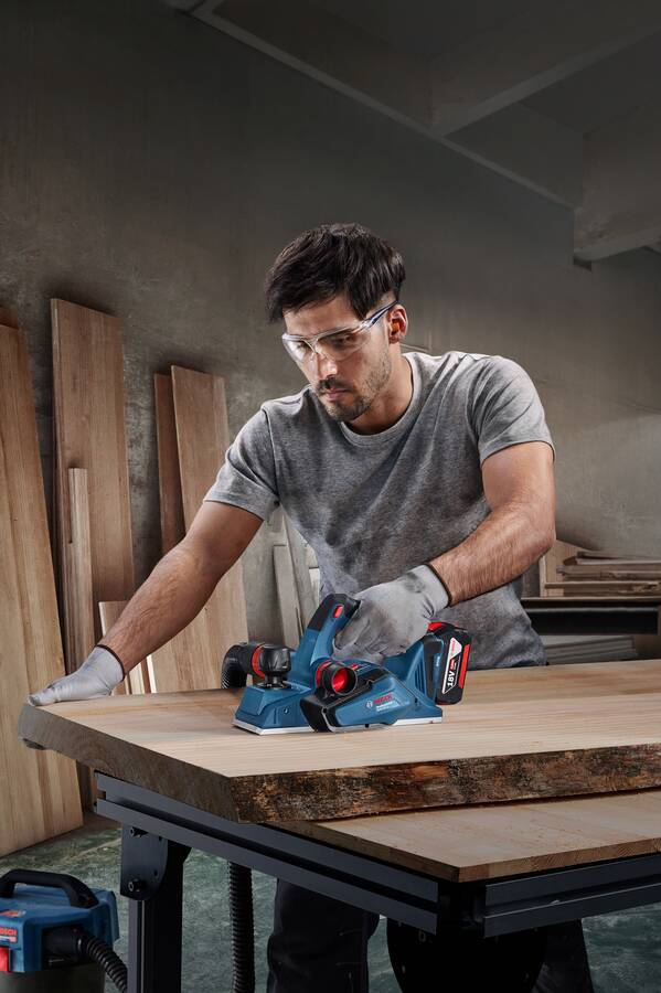 Eine Person mit Sicherheitsbrille verwendet einen blauen handgehaltenen Elektrohobel auf einem Holzbrett und konzentriert sich darauf, die Oberfläche zu glätten.