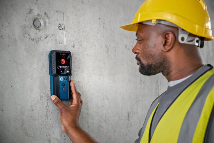 Ein Bauarbeiter mit einem gelben Schutzhelm verwendet einen Wandscanner, um Objekte innerhalb einer Betonwand zu erkennen.