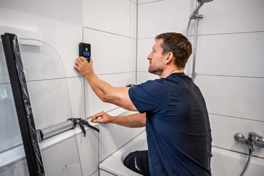 Ein Mann kniet in einem Badezimmer neben einer Badewanne und justiert einen an der Wand befestigten Audiomonitor.