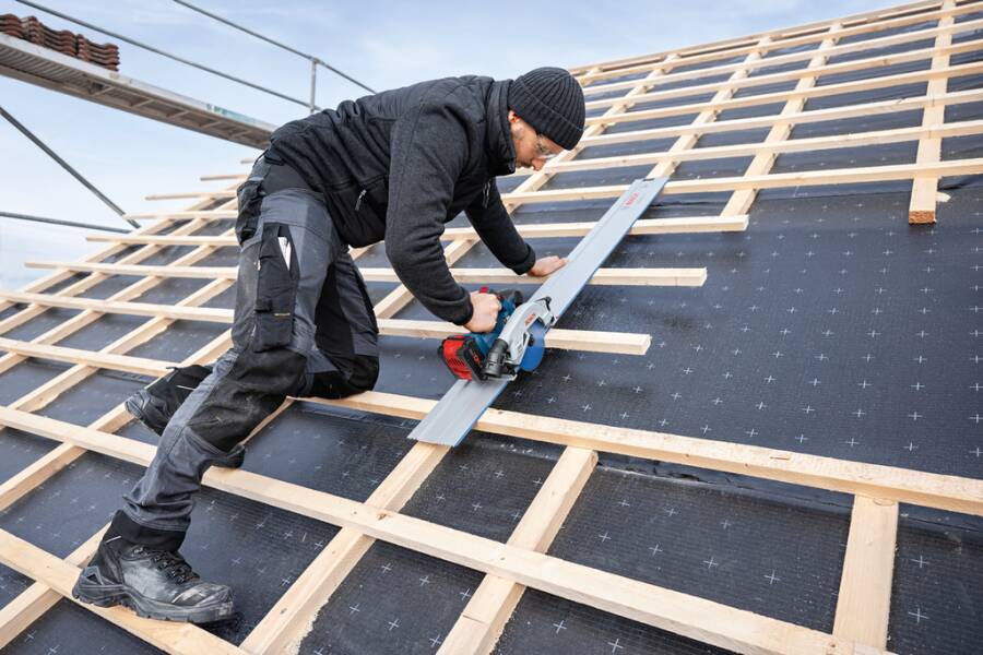 Arbeiter, der Dachbaumaterial auf einem geneigten Dach mit einer Kreissäge schneidet und dabei Schutzkleidung und Schutzausrüstung trägt. Dachstuhl und Himmel sind sichtbar.
