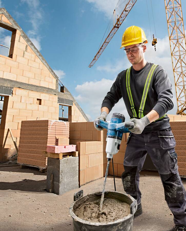 Ein Bauarbeiter mit Schutzhelm und Warnweste bedient einen Betonmischer auf einer Baustelle mit Ziegelmauern und einem Kran im Hintergrund.